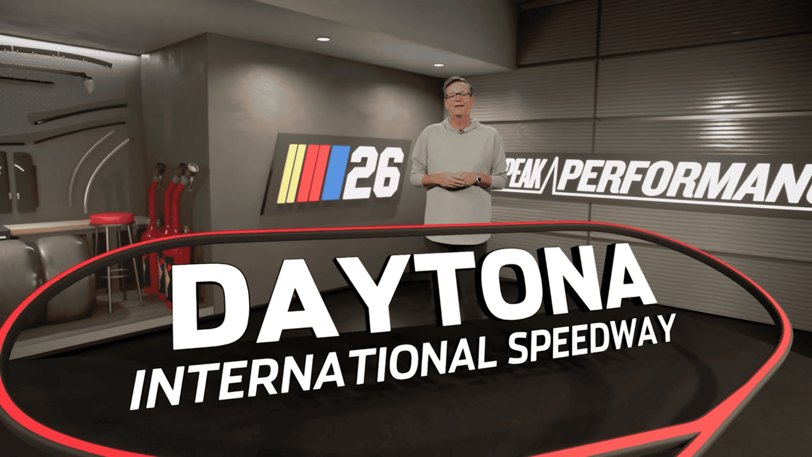 NASCAR 26 promotional screenshot showing a host presenting Daytona International Speedway in a Peak Performance garage studio setting with the NASCAR 26 logo
